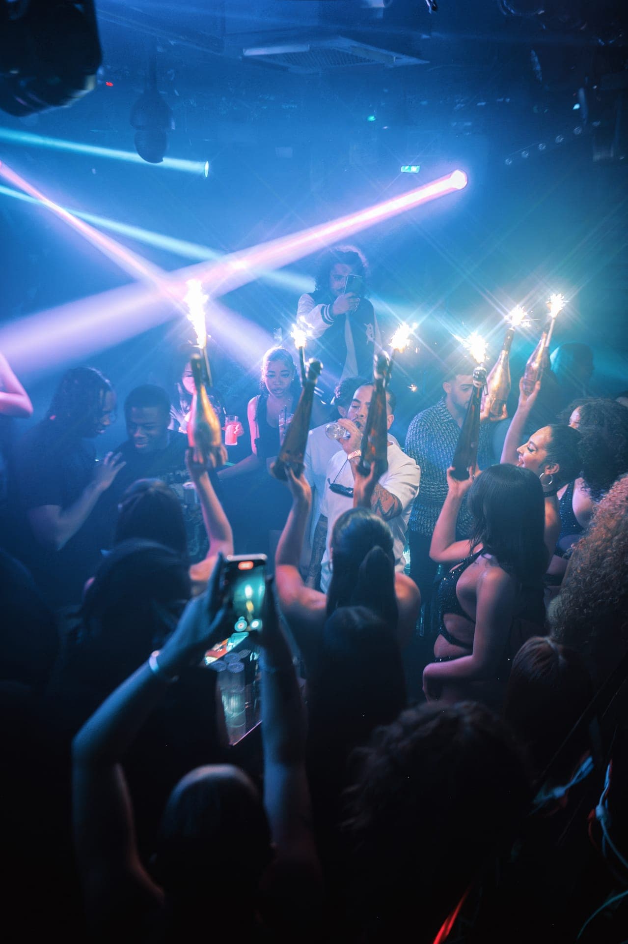 A crowded nightclub dance floor with guests holding sparkler-topped bottles under blue and pink lights