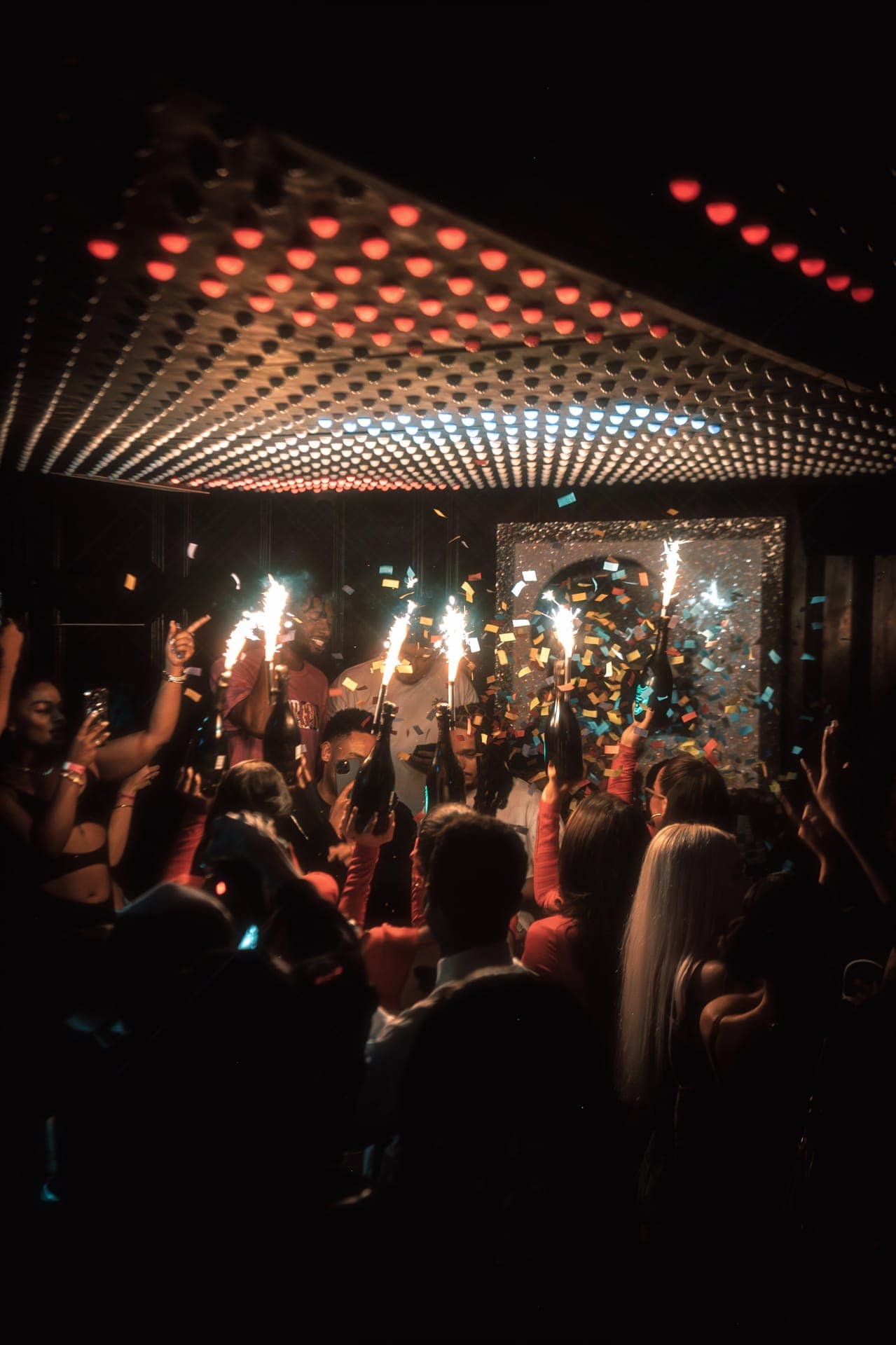 Crowd raising sparkler-topped bottles under a dotted light ceiling as confetti falls
