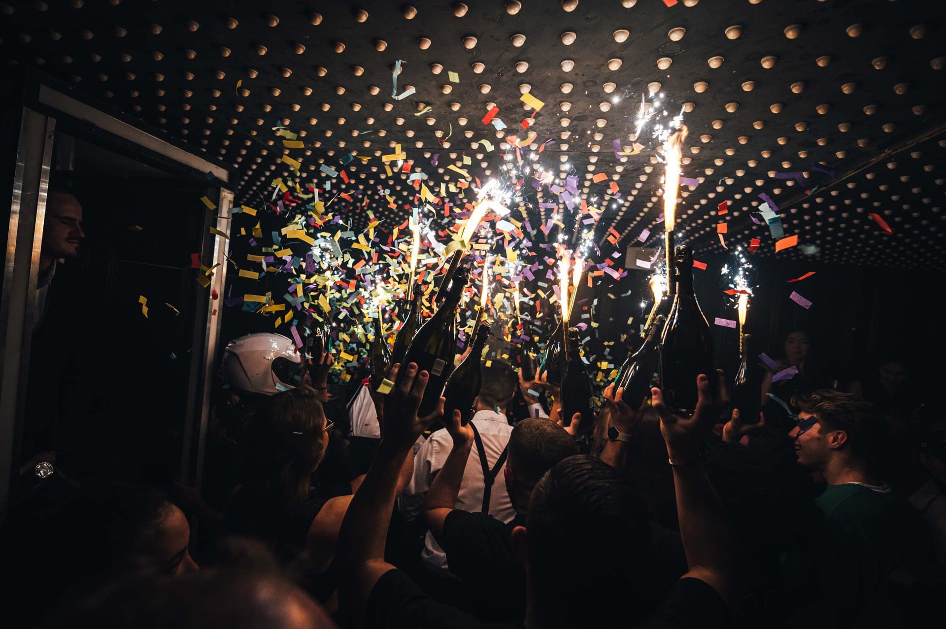 Champagne bottles with sparklers held up under falling confetti in a crowded nightclub