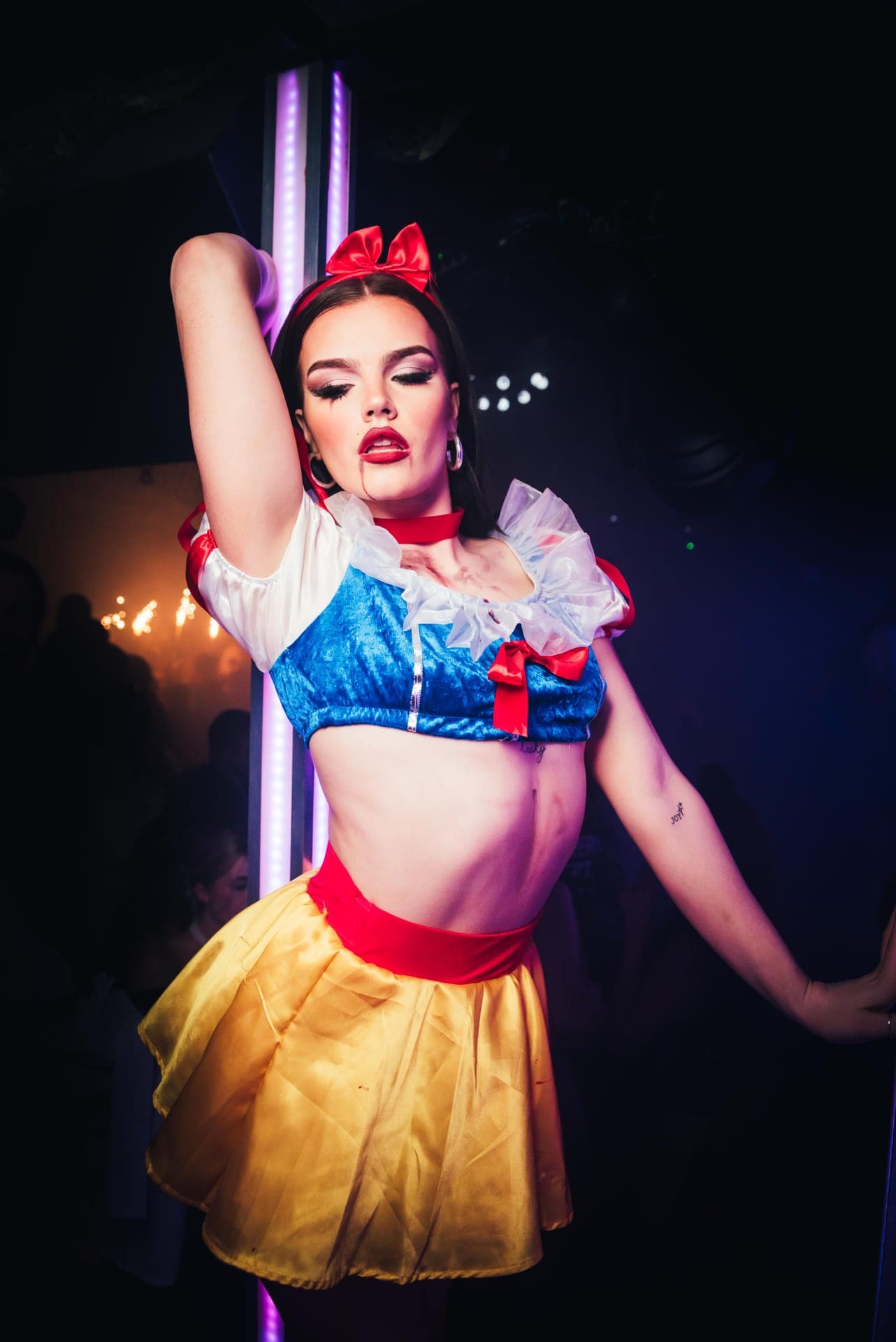 A woman in a Snow White Halloween costume with fake blood at her lip posing beside a purple-lit pole in a dark club